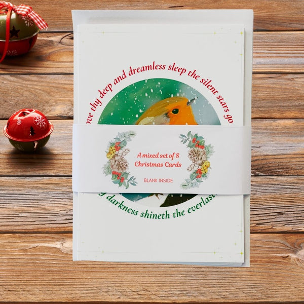 A mixed set of 8 Mary, Martha & Meg Christmas Greetings cards. The card shown here is of a winter robin framed with lyrics from the traditional Christmas carol, "O Little Town of Bethlehem." The pack of 8 cards and envelopes is held together by a belly band which features the words, A mixed set of 8 Christmas Cards. Either side of this is a wreath motif featured on some of the cards included in the pack. This beautiful set of cards is photographed on a wooden table with Christmas baubles.