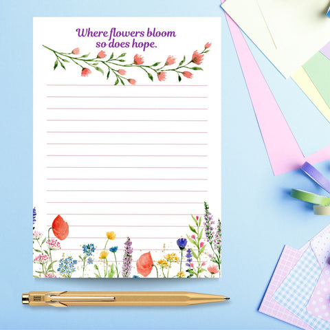 A smooth, white notepad with pink lines and delicate watercolour wildflowers. The text at the top of the page reads, ‘Where flowers bloom so does hope.’ Underneath this is stem of pretty pink flowers and green leaves. The lower part of the notepad is covered in lovely flowers of different colours. This Mary, Martha & Meg notepad is pictured on a pale blue background with pastel stationary items to the right and a gold pen underneath the notepad. A lovely gift to help you stay organised.