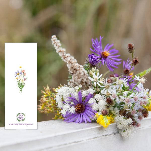 The reverse side of a Mary, Martha & Meg ‘’Send Me’ bookmark. The white background of the card features is brought to life with a miniature version of the wildflower bouquet on the front of the bookmark. At the bottom of the bookmark is the Mary, Martha & Meg logo, a green wreath with tiny purple flowers. Underneath this is a website reference: www.marymarthameg.co.uk The bookmark is placed on a windowsill next to a bunch of flowers.