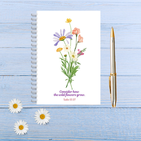 A pretty white notepad with a white spiral binding. At the centre of the notepad is a bunch of beautiful wildflowers and delicate green foliage. The Bible verse reads, “Consider how the wildflowers grow.” Luke 12:27. The notebook is pictured on a blue wooden table, with a silver and gold pen to the right of the notebook, and three daisies in the right-hand corner. This practical Mary, Martha & Meg notebook is A6 sized – perfect for popping into a handbag.