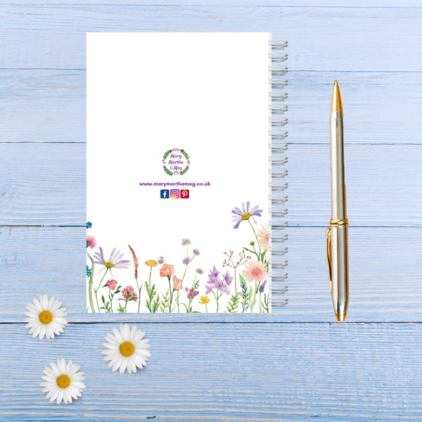 The back cover of a pretty white notepad with a white spiral binding. At the centre is the Mary, Martha & Meg logo of green leaves and tiny purple flowers. Underneath are small social media logos of where Mary, Martha & Meg products can be found. The eye is drawn to the beautiful wildflowers. The notebook is pictured on a blue wooden table, with a silver and gold pen to the right of the notebook, and three daisies in the right-hand corner.