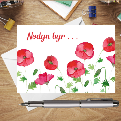 A classic Mary, Martha & Meg postcard, but in Welsh! The digital text, Nodyn Byr means ‘little note’ in Welsh. The postcard features beautiful poppies which have been hand-painted using watercolours. The edge of a white envelope can be seen behind the postcard. The postcard is placed on a wooden writing surface with a silver pen beneath it and stationary items above it. A wonderful Welsh postcard to help you stay in touch with your friends.