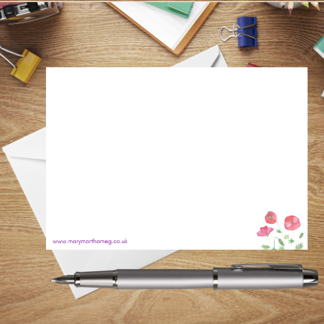 A photograph showing the reverse of a Nodyn Byr postcard. It features a small red poppy motif in the lower right corner, as well as a small reference to the Mary, Martha & Meg website: www.marymarthameg.co.uk . The edge of a white envelope can be seen behind the postcard. The postcard is placed on a wooden writing surface with a silver pen and stationary items.