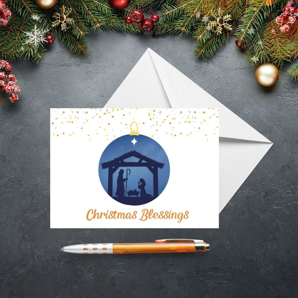 This Mary, Martha & Meg Christmas Greetings card features a Nativity scene showing the silhouettes of Mary and Joseph in a stable, worshipping baby Jesus lying in a manger. The scene is captured inside a Christmas bauble of an inky blue sky. Golden stars sit above the bauble. The greeting reads, ‘Christmas Blessings.’ The card is photographed on a slate gray background with Christmas fir tree branches and festive decorations.