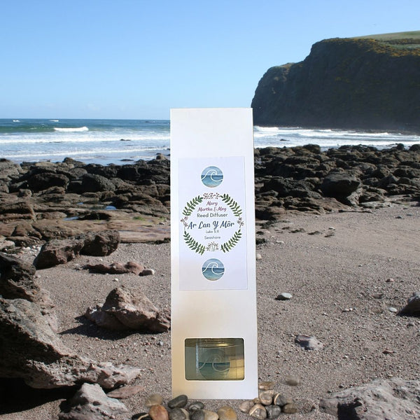 A picture showing a Mary, Martha & Meg luxury reed diffuser inside its white packaging box. The box shows a Mary, Martha & Meg logo. Inside the logo can be seen ‘Ar Lan Y Môr’ the Welsh words for ‘By the seashore.’ A small image of a blue wave is above and below the logo design. The small rectangle cut-out in the box shows the glass bottle of the reed diffuser. The box is positioned on a rocky seashore.