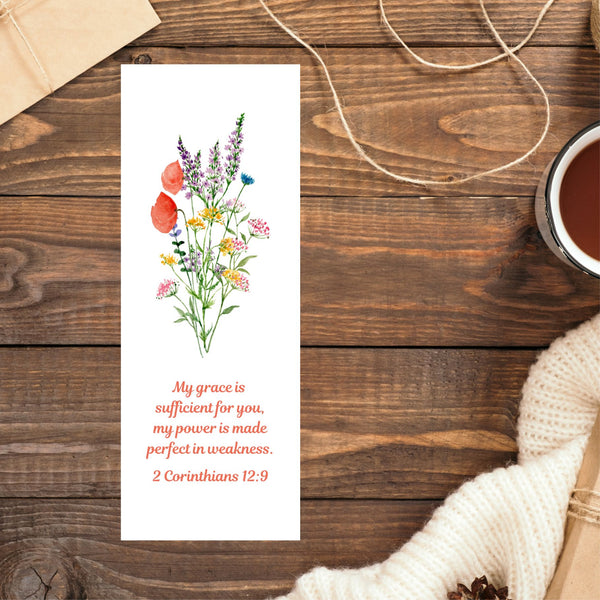 A beautiful Mary, Martha & Meg bookmark featuring a pretty wildflower floral bouquet. Underneath the floral design is the encouraging Bible verse, โMy grace is sufficient for you, my power is made perfect in weakness.โ 2 Corinthians 12:9. The bookmark is pictured on a brown wooden table. In the top left corner the edge of a brown parcel can be seen. In the top right corner there is some loose string. A cup of coffee and a cream jumper can be seen on the right side of the photo.