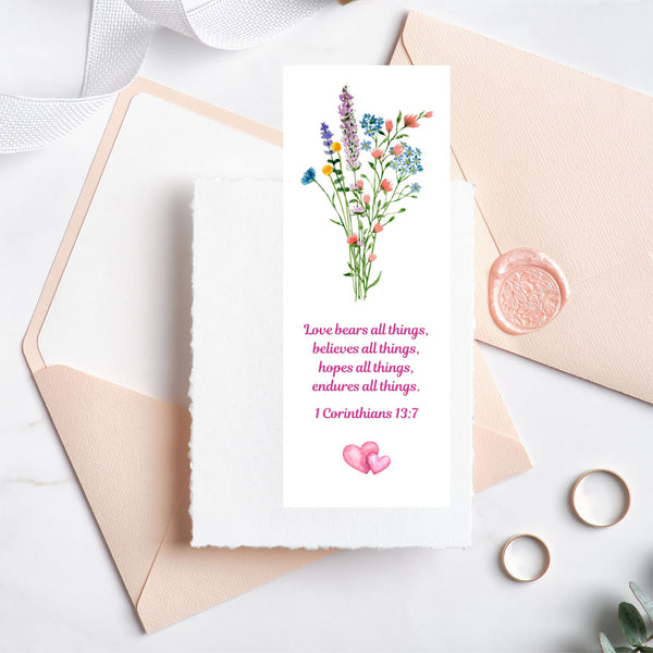 A Mary, Martha & Meg bookmark featuring a beautiful wildflower floral bouquet. Underneath this is the treasured verse, “Love bears all things, believes all things, hopes all things, endures all things.” 1 Corinthians 13:7. Two pink intertwined hearts are below the Bible verse. The bookmark is pictured on top of cream and peach stationary on a white background. Two wedding bands are place in the lower right corner.