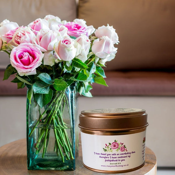 A photograph of a Mary, Martha & Meg small soy wax scented candle placed next to a vase of roses. This picture shows the reverse of the Cariad candle label. The purple text is “I have loved you with an everlasting love; therefore I have continued my faithfulness to you,” Jeremiah 31:3. Above the Bible verse is a small design of pink roses, taken from an original watercolour painting by Mary, Martha & Meg. The candle tin is rose gold. A perfect for a gift for a loved one.