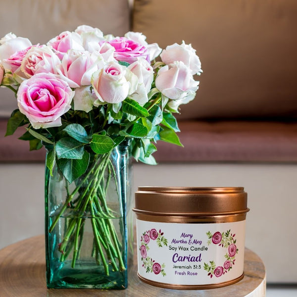 A photograph of a Mary, Martha & Meg small soy wax scented candle placed next to a vase of roses. The label of this natural vegan soy candle shows the name of the candle, Cariad, which is the Welsh word for love. There is a reference to the inspiration for this beautiful candle, Jeremiah 31:3. The white label is framed by pink roses. The tin is rose gold. A perfect for a gift for a loved one.