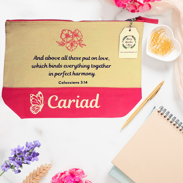 A natural canvas zip pouch with a pink zip and pink base. The Bible verse reads, "And above all these put on love, which binds everything together in perfect harmony." Colossians 3:14. Above the text is a trio of pink flowers and petite leaves. A Mary, Martha & Meg tag can be seen hanging from the zipper on the right of the pouch. The pink base of the pouch has the Welsh word 'Cariad' (meaning love) and a beautiful butterfly.This beautiful item is pictured on on a table with stationary items.