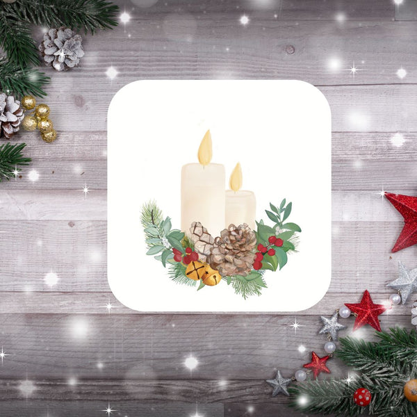 A festive Christmas coaster created by Mary, Martha & Meg. The square coaster has rounded edges. The design is of two lit candles resting on a Christmas wreath of holly berries, pine cones, Christmas bells and green foliage. This pretty Christmas coaster is pictured on a pale grey background with shining lights. Christmas decorations peep from the top right and bottom left corners of the photograph.
