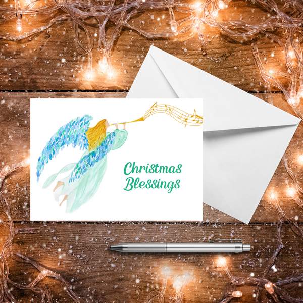 A Mary, Martha & Meg Christmas Greeting card featuring a heralding angel, from an original Mary, Martha & Meg watercolour. The golden-haired angel wears a delicate mint gown and has beautiful blue and green feathered wings. Musical notes flow from the trumpet she is blowing. The text reads, ‘Christmas Blessings.’ A white envelope sits behind the card.  These are photographed on a wooden table with warm Christmas lights and a silver pen.