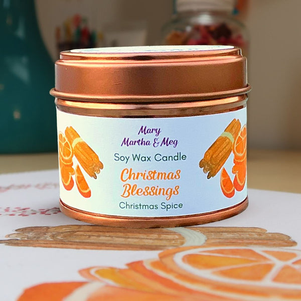 A Mary, Martha & Meg Christmas Blessing soy wax candle. The candle fragrance is Christmas Spice, a delightful seasonal blend of spices and fruit associated with Christmas. The Mary, Martha & Meg label shows hand-painted slices of oranges and cinnamon sticks. The candle  is sitting on part of the original watercolour painting for the label design.