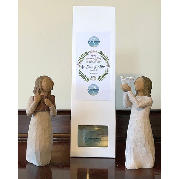 A photograph showing a Mary, Martha and Meg ‘Seashore’ Reed Diffuser in its white packaging. The front of the white packaging box has the Mary, Martha & Meg logo. Inside this is the name of the fragrance, ‘Ar Lan Y Môr,’ which translates into English as ‘By the Seashore.’ Above and below the logo are images of blue waves. The diffuser bottle can be seen through the rectangular cut-out in the box. Two wooden figurines have been placed either side of the product, for illustration purposes only.