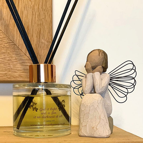 A Mary, Martha & Meg luxury reed diffuser bottle with a rose gold coloured top is seen placed on a wooden mantlepiece below a wooden-framed mirror. The Bible verse which inspired this Black Orchid ‘Hope’ fragrance can be seen, “God is Light and in Him is no darkness at all.” 1 John 1:5. Below this is a reference to Mary, Martha & Meg website. To the right of the diffuser is a wooden sitting angel.