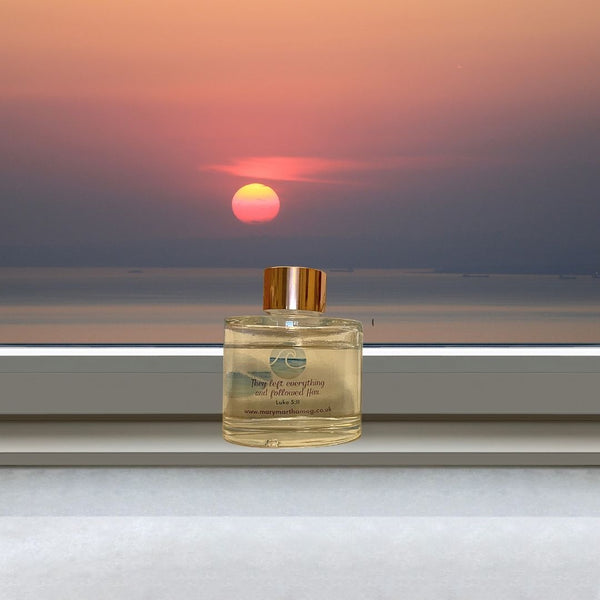 A Mary, Martha & Meg luxury reed diffuser bottle with a gold-coloured top is seen placed on a windowsill with a view of the sun setting over the sea in the background. The Bible verse which inspired this seashore fragrance, ‘They left everything and followed Him,’ Luke 5:11, is included on the bottle, as well as a reference to Mary, Martha & Meg website.
