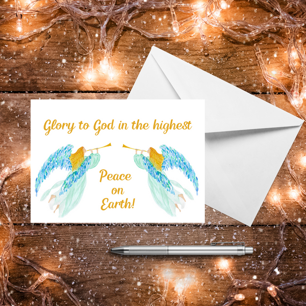 A Mary, Martha & Meg Christmas Greeting card featuring two heralding angels. The golden-haired angels wear delicate mint gowns and have beautiful blue and green feathered wings. Each carries a golden trumpet. The text reads, ‘Glory to God in the highest, peace on Earth!’ This is from the Bibles verse found in Luke 2:14. A white envelope A white envelope sits behind the card. These are photographed on a wooden table with warm Christmas lights and a silver pen.