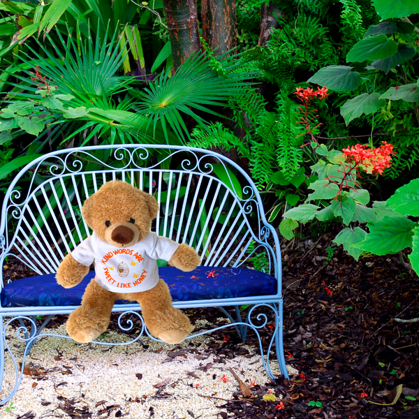 A photo of a Mary, Martha & Meg plush Honey Bear sitting on a miniature blue bench in a woodland setting of green leaves, orange flowers, tree bark. This beautifully soft bear wears a white t-shirt with the hand-printed orange text, "Kind words are sweet like honey." Honey Bear is a wonderful reminder of God's encouragement to be kind and show love to others.