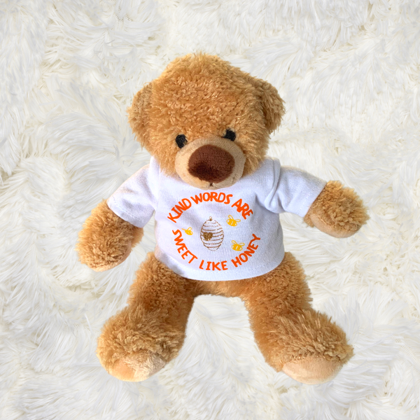A photograph of a Mary, Martha & Meg Honey Bear ‘Kind Words’ soft toy, which is pictured resting on a white pillow. The soft to touch fur teddy bear has a friendly face and wears a t-shirt with the words from Proverbs 16:24, "Kind words are sweet like honey." The orange text words are hand-printed in a circular shape. Inside the words is a golden glitter honeycomb with a gold glitter heart at its centre. Three yellow bees dance around the honeycomb.