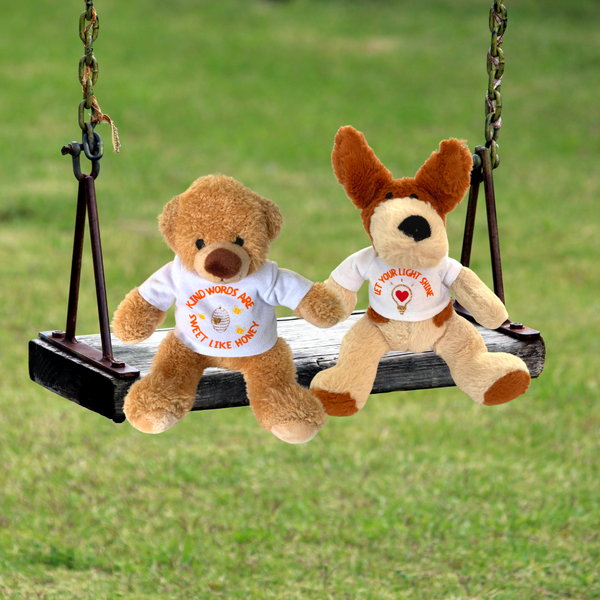 A photograph of two of Mary, Martha & Meg delightful soft toys – Honey Bear and the ‘pawsome’ Let your Light Shine cute dog. The soft plush toys are both wearing white t-shirts with encouraging words. Honey Bear’s t-shirt reads, "Kind words are sweet like honey." The soft pawsome dog wears a t-shirt which says, ’Let your light shine.’ Both soft toys are holding paws, seated on a miniature swing on a green lawn. Great gifts of Christian encouragement.
