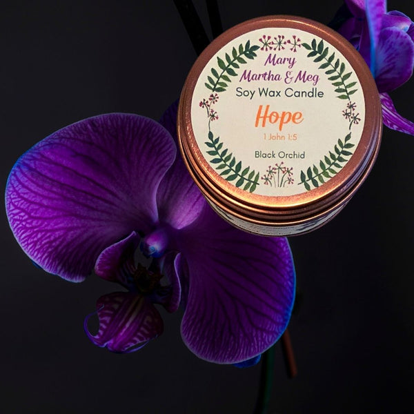 This photograph shows the rose gold lid of the Mary, Martha & Meg soy wax candle named ‘Hope.’ There is a small reference to 1 John 1:5, which refers to the inspiration for the candle. Underneath this is the name of this luxurious fragrance – Black Orchid, a gorgeous harmony of ylang, bergamot, truffle, orchid, dark chocolate and more! This small soy wax candle is pictured on a black background with a dark purple orchid, matching the Mary, Martha & Meg logo perfectly.