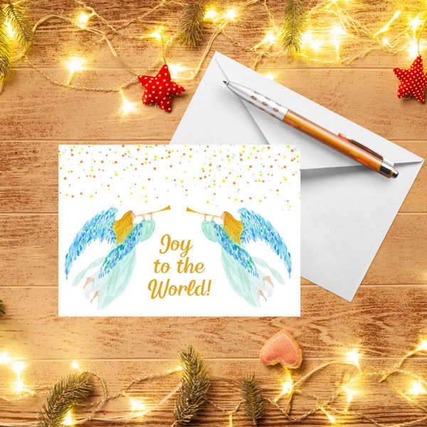 A Mary, Martha & Meg Christmas Greeting card featuring two heralding angels. The golden-haired angels wear delicate mint gowns and have beautiful blue and green feathered wings. Each carries a golden trumpet. Above them shine tiny stars. The text reads, ‘Joy to the World.’ A white envelope and pen are behind the card. These are photographed on a wooden table with warm Christmas lights and red handmade decorations.