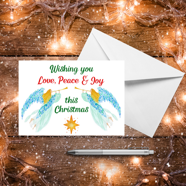 A Mary, Martha & Meg Christmas Greeting card featuring two heralding angels. The golden-haired angels wear delicate mint gowns and have beautiful blue and green feathered wings. Each carries a golden trumpet. The text reads, ‘Wishing you Peace & Joy this Christmas.’ There is a golden star underneath the greeting. A white envelope sits behind the card. These are photographed on a wooden table with warm Christmas lights and a silver pen.