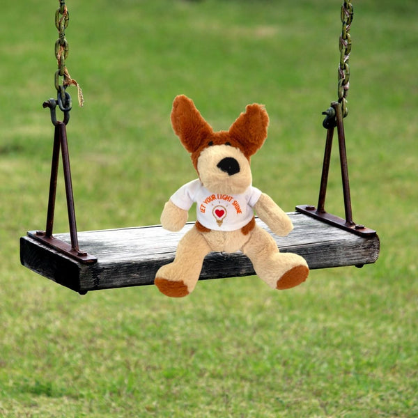 A totally ‘pawsome’ Mary, Martha & Meg soft toy dog. This small plush pup is cream with light brown ears and features. He is wearing a white t-shirt with the words, 'Let your light shine.' This positive encouragement can be given to any child, including those who are still young at heart. The orange text is curved above a design of a gold glitter lightbulb with a red heart at its centre. This adorable soft toy dog is seated on a swing seat with green grass in the background.