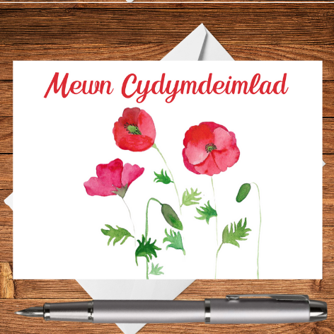 A Mary, Martha & Meg card lovingly created for someone going through a time of loss. The card features a design of three delicately hand-painted red poppies. Mewn Cydymdeimlad is digitally added above the floral design. The edges of a crisp white envelope can be seen behind the card, which rests on a wooden writing surface. A silver pen has been placed beneath the sympathy card. A comforting card to let someone know that you are thinking of them at such a difficult time.