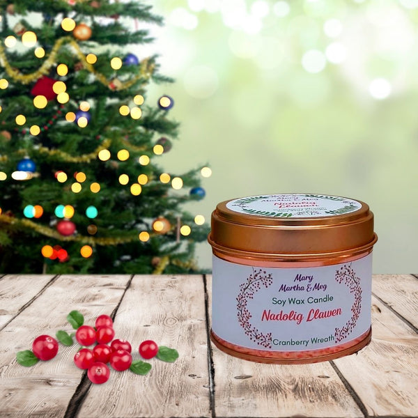 A small Mary, Martha & Meg natural soy wax candle. Red text shows the name of the candle, ‘Nadolig Llawen.’ Underneath, in green text, is the candle fragrance, ‘Cranberry Wreath.’ The centred text is framed by a hand-painted image of a cranberry wreath. The candle tin and lid are rose gold. The candle tin sits on wood. To the right of the candle are cranberries. There is a brightly lit Christmas tree in the background.