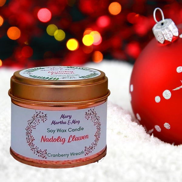 A Mary, Martha & Meg natural soy wax candle. Red text shows the name of the candle, ‘Nadolig Llawen.’ Underneath, in green text, is the candle fragrance, ‘Cranberry Wreath.’ The centred text is framed by a hand-painted image of a cranberry wreath. The candle tin and lid are rose gold. The candle tin sits on snow, with a red and white bauble to the right. The background is red with yellow and orange coloured lights.
