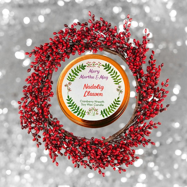 A Mary, Martha & Meg rose gold candle lid. The white label has the name of the candle, Nadolig Llawen, in red text in the centre of the label. Underneath, in green text, is the name of the fragrance, Cranberry Wreath, as well as the text ‘Soy Wax Candle.’ All text is framed by the Mary, Martha and Meg logo of a green wreath of leaves with tiny flowers. The candle lid sits inside a wreath of red cranberries. Behind this is a silver background.