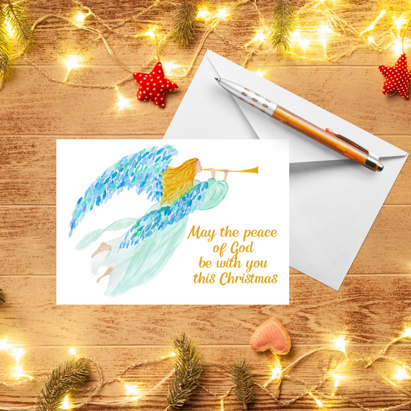 A Mary, Martha & Meg Christmas Greeting card featuring a heralding angel, from an original Mary, Martha & Meg watercolour. The golden-haired angel wears a delicate mint gown and has beautiful blue and green feathered wings. She carries a trumpet. The text reads, ‘May the peace of God be with you this Christmas.’ A white envelope and pen are behind the card. These are photographed on a wooden table with warm Christmas lights and red handmade decorations.