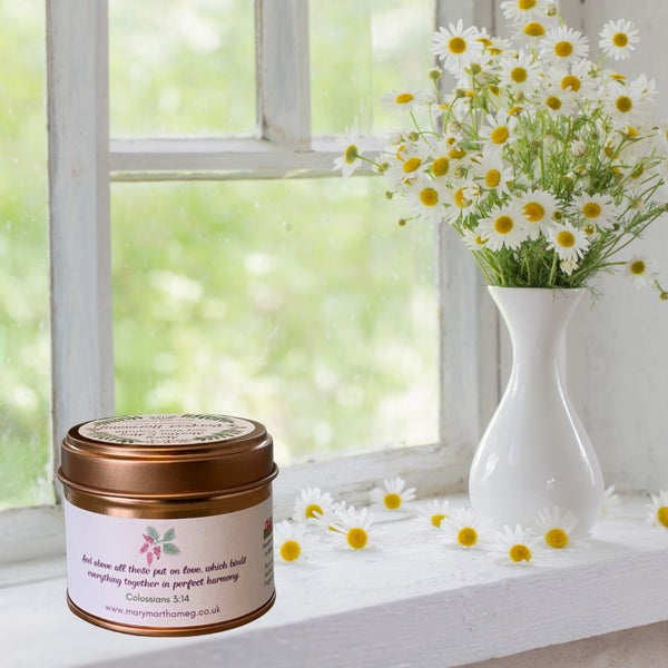 A Mary, Martha & Meg soy wax scented small candle tin pictured on a white windowsill next to a vase of daisies, with individual daisies scattered next to the candle. The photo shows the reverse of the candle label Perfect Harmony, which reads, “And above all these put on love, which binds everything together in perfect harmony.” Colossians 3:14. The natural soy wax is fragranced with the beautiful floral notes of plum and mulberry.