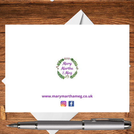 A photograph showing the reverse of a Nodyn Byr greetings card. It features a small red poppy motif in the lower right corner, as well as a small reference to the Mary, Martha & Meg website: www.marymarthameg.co.uk . The edge of a white envelope can be seen behind the postcard. The postcard is placed on a wooden writing surface with a silver pen.