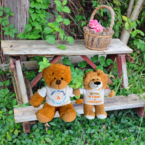 A photograph of two of Mary, Martha & Meg delightful soft toys – Prayer Teddy Bear and Louis the small lion. The soft plush toys are both wearing white t-shirts showing Bible verses. Prayer Teddy Bear has the hand-printed verse, ‘When I am afraid I will trust in you,’ whilst Louis the Lion has the words ‘Be strong and courageous.’ They are seated on a small wooden picnic bench with a wicker basket of pink flowers on the table, which is surrounded by green leaves.