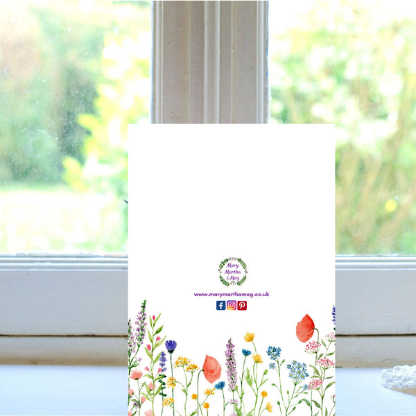 The reverse side of a Mary, Martha & Meg ‘Rejoice’ card. The white background of the card features delicate flowers and is a lovely addition to the card, adding a touch of elegance to the card. At the centre of the card is the Mary, Martha & Meg logo. Underneath are references to the Mary, Martha & Meg website and social media sites. The card is pictured on a white windowsill.