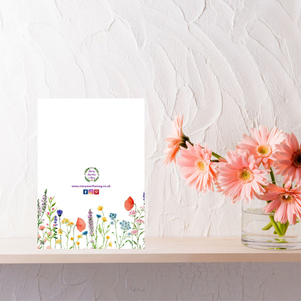 The reverse side of a Mary, Martha & Meg ‘Praying for You’ card. The white background of the card features delicate flowers and is a lovely addition to the card, adding a touch of elegance to the card. At the centre of the card is the Mary, Martha & Meg logo. Underneath are references to the Mary, Martha & Meg website and social media sites. The card is pictured on a pale brown shelf next to a vase of peach flowers.