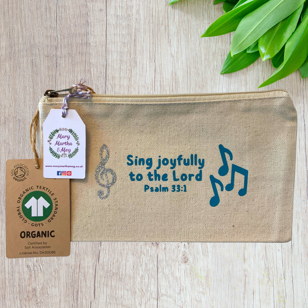 A Mary, Martha & Meg organic, natural, canvas zip pouch featuring the Bible verse, ‘Sing joyfully to the Lord. Psalm 33:1.’ The design also features a silver glitter treble clef and blue musical notes. The design has been hand-printed. A Mary, Martha & Meg tag hangs from the zip, as well as a label showing the organic credentials of the zip pouch. The zip pouch is pictured on a wooden table with green leaves in the top right corner. A great, practical gift which can be posted through a letterbox.