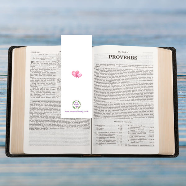 The reverse of a Mary, Martha & Meg ‘Rejoice’ bookmark. Two pink intertwined hearts are in the centre of the white bookmark. The Mary, Martha & Meg logo of a green wreath with tiny purple flowers is featured near the bottom of the bookmark. Underneath this is a reference to the website marymarthameg.co.uk . The bookmark is photographed in a Bible open to the book of Proverbs. The Bible rests on a pale blue background.