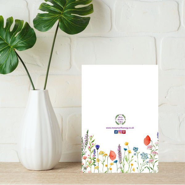 The reverse side of a Mary, Martha & Meg ‘Rejoice card. The white background of the card features delicate flowers and is a lovely addition to the card, adding a touch of elegance to the card. At the centre of the card is the Mary, Martha & Meg logo. Underneath are references to the Mary, Martha & Meg website and social media sites. The card is pictured on a pale brown shelf with white painted bricks in the background. Next to the card is a white vase with bold green leaves.