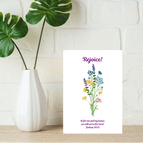 A Mary Martha & Meg A6 white card featuring the word ‘Rejoice’ above a floral bouquet of pretty wildflowers. Underneath the flowers is the Bible verse, “As for me and my house, we will serve the Lord.” Joshua 24:15. The card is pictured on a pale brown shelf with white painted bricks in the background. Next to the card is a white vase with bold green leaves.