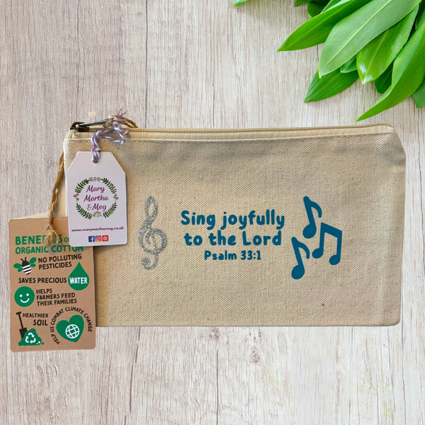 A Mary, Martha & Meg organic, natural, canvas zip pouch featuring the Bible verse, ‘Sing joyfully to the Lord. Psalm 33:1.’ The design also features a silver glitter treble clef and blue musical notes. The design has been hand-printed. A Mary, Martha & Meg tag hangs from the zip. The zip pouch label shows the reverse of the label in the previous photograph, illustrating the benefits of organic cotton. The zip pouch is pictured on a wooden table with green leaves in the top right corner.