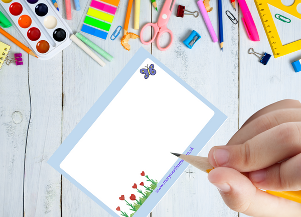 This photograph shows the reverse of the ‘You are amazing’ postcard. The reverse is brightened by smaller images from the front of the postcard: there is a small purple and yellow butterfly in the top right corner. In the lower corner there are small flowers with red hearts growing among green grass. There is a small reference to the Mary, Martha & Meg website at the bottom of the postcard. A child’s hand is seen with a pencil in hand. The postcard lies on a white wooden table with stationary items.