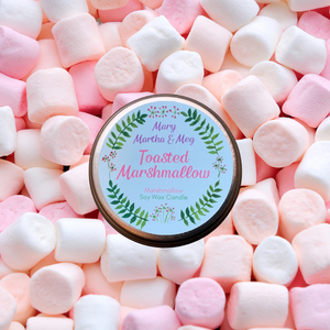A photograph showing the lid of a small Mary, Martha & Meg 'Toasted Marshmallow' candle. The rose-rimmed lid has a Mary, Martha & Meg sticker showing the  name of the candle in pink lettering, inside a wreath of green leaves. The lid has been pictured in a bed of pink and peach marshmallows.