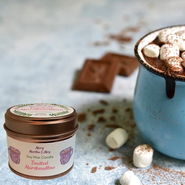 A small Mary, Martha & Meg soy wax candle pictured next to a blue mug of hot chocolate and marshmallows. On the lace table cloth there are also three white marshmallows and two pieces of square chocolate. The white label on the rose gold candle tin shows the name of its maker - Mary, Martha & Meg. The words 'soy wax candle' are included as well as the name of the fragrance, 'Toasted Marshmallow.' There are 2 purple cups of hot chocolate either side of the text on the label.