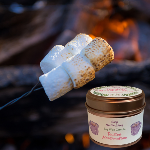 A small Mary, Martha & Meg soy wax candle in a rose gold tin. The name of the candle, 'Toasted Marshmallow,' is written in pink font on the white label. There are two purple cups of cocoa either side of the text. The candle is pictured to the left of toasted marshmallows on a metal toasting stick, with a blurred fire in the background.
