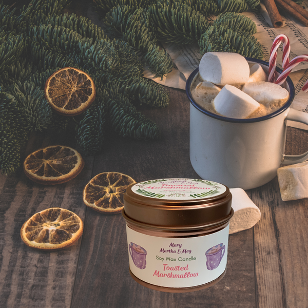 A small Mary, Martha & Meg candle pictured on a brown wooden table. To the left of the candle are slices of dried oranges. To the right is a mug of hot chocolate topped with white marshmallows and two red and white candy canes. Green pine leaves are seen in the background. Peeping through the top right corner of the picture are pages of music and cinnamon sticks.