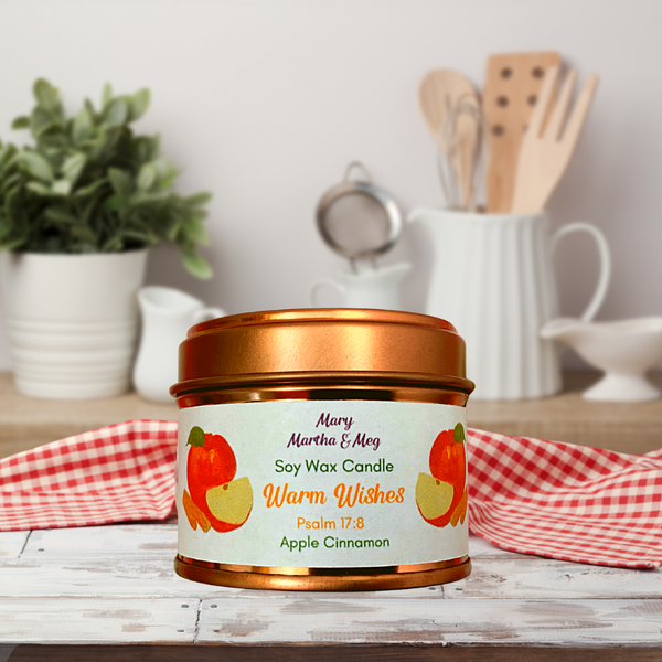'Warm Wishes' Apple Cinnamon Soy wax candle in a rose gold tin. A wonderful Baked Apple and warm spice fragrance. The background is a kitchen with utensils and plant. This Mary, Martha & Meg soy wax candle comes in a rose gold tin. The front of the tin has the candle fragrance name which is 'Warm Wishes' and accompanied with a hand-painted juicy red apple and cinnamon sticks.