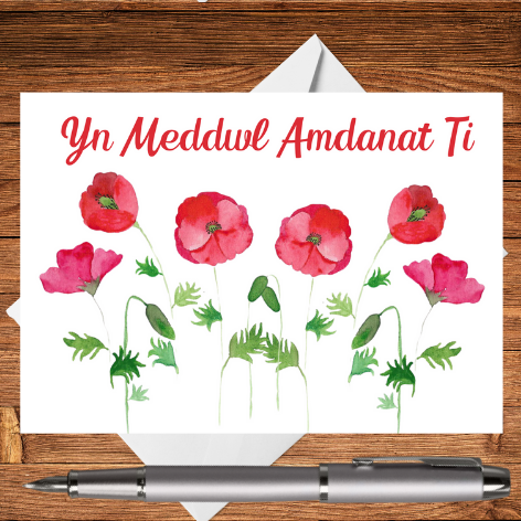 A classic Mary, Martha & Meg thinking of you greetings card in Welsh – Yn Meddwl Amdanat Ti. This cerdyn cyfarch features red poppies (pabïau goch), which have been hand-painted using watercolours. The edges of a crisp white envelope can be seen behind the card, which rests on a wooden writing surface. A silver pen has been placed beneath the card. A perfect card to remind a Welsh friend you are thinking of them.
